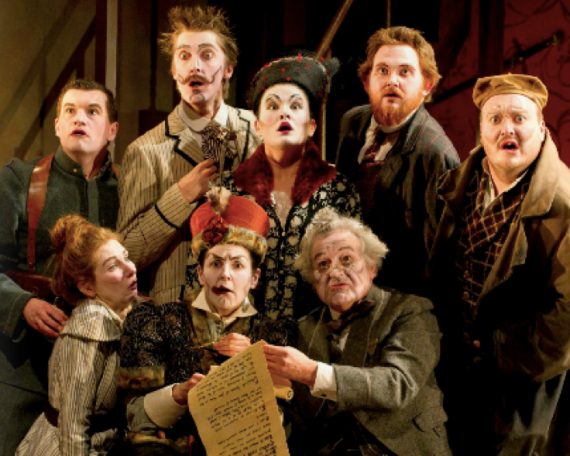 An opera production image. A group of characters look surprised as a will is read out